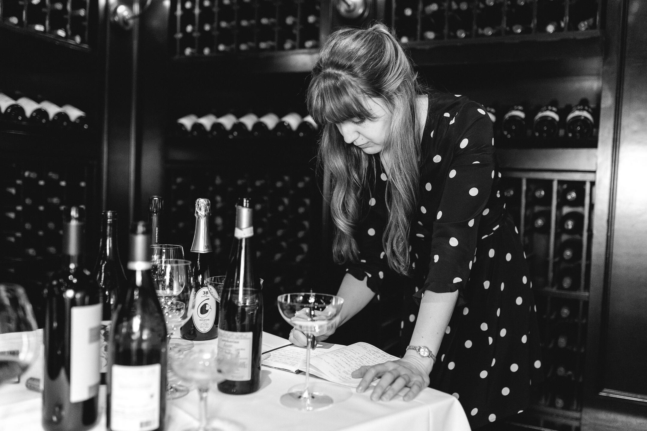 Studying Wine A woman wearing a polka-dot dress, standing over a table full of wine bottles and glasses, writing in a journal